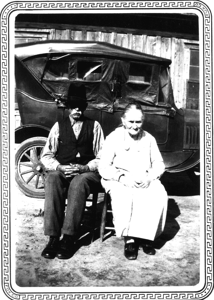 A black and white photo of a man and woman sitting in front of an old vehicle